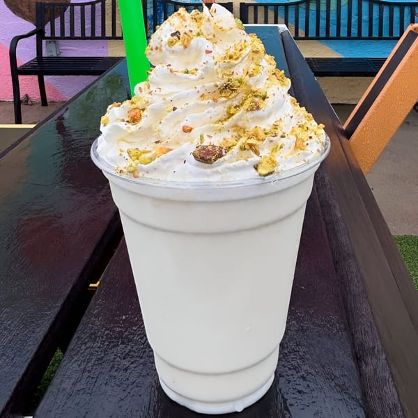 A white milkshake topped with whipped cream and crushed nuts sits on a dark outdoor table.