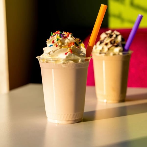 Two milkshakes with whipped cream and sprinkles, one with an orange straw, the other with a purple straw.