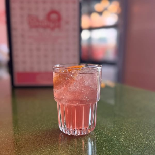 Pink cocktail with ice and an orange slice on a green glittery table at Baby's.