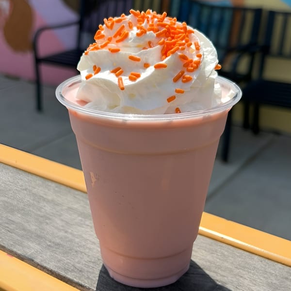 Pink milkshake with whipped cream and orange sprinkles on a wooden table outdoors.